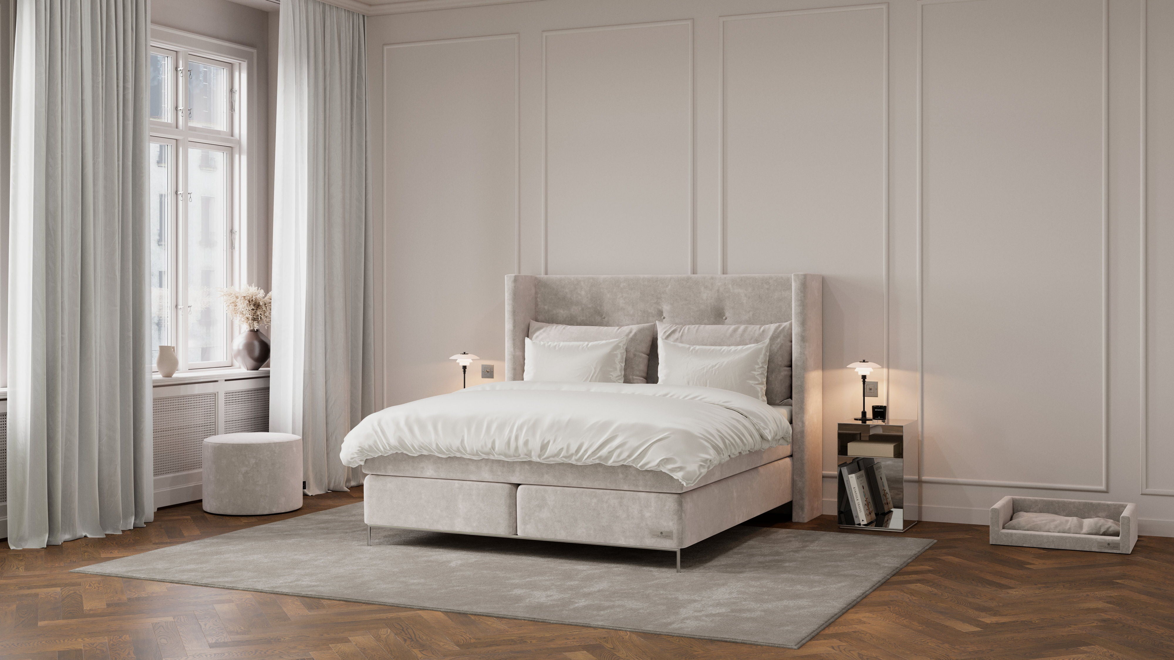 A modern bedroom featuring a light gray upholstered bed with white bedding, set against a beige paneled wall with a window, curtains, and a gray rug.